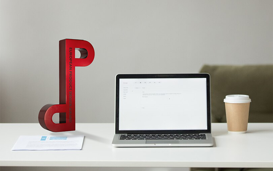 Trofeo dell’European Product Design Award in metallo spazzolato rosso su una scrivania da ufficio, con l’ambiente visibile attraverso le aperture dei suoi occhielli